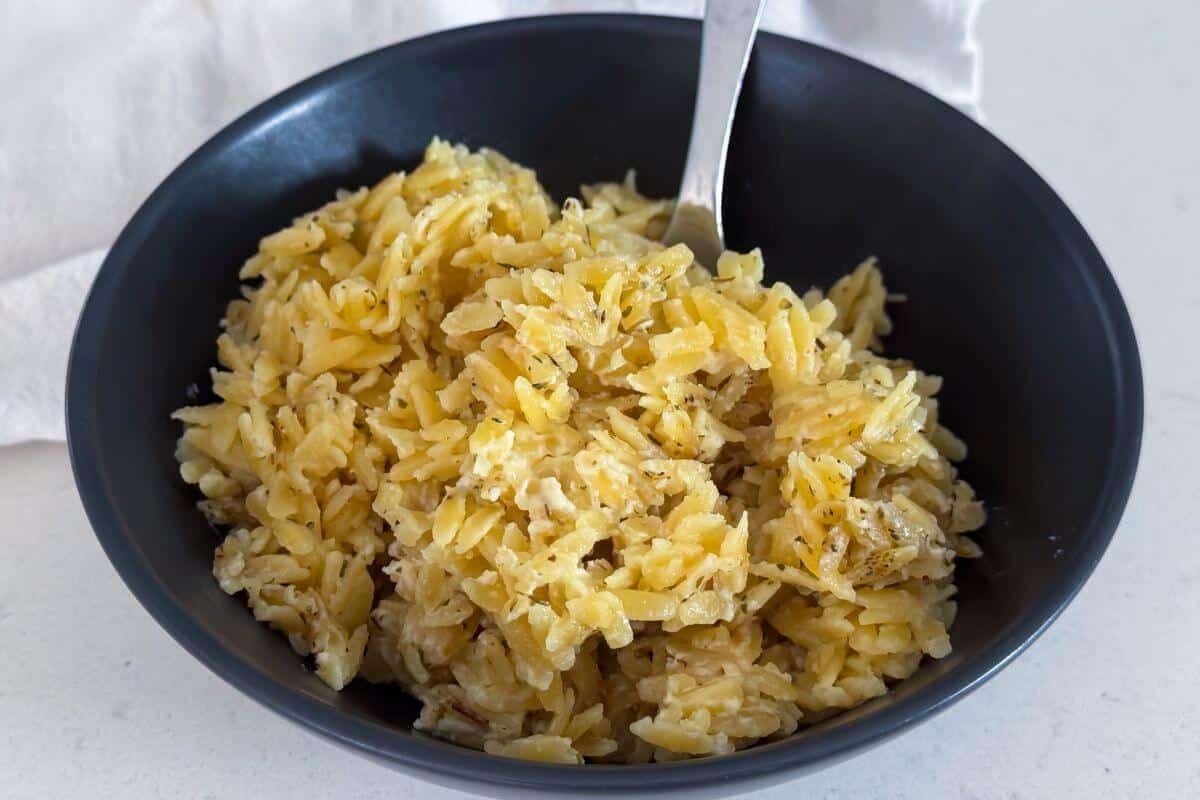 A black bowl filled with cooked orzo pasta in a creamy, yellow sauce, with a silver spoon resting inside.