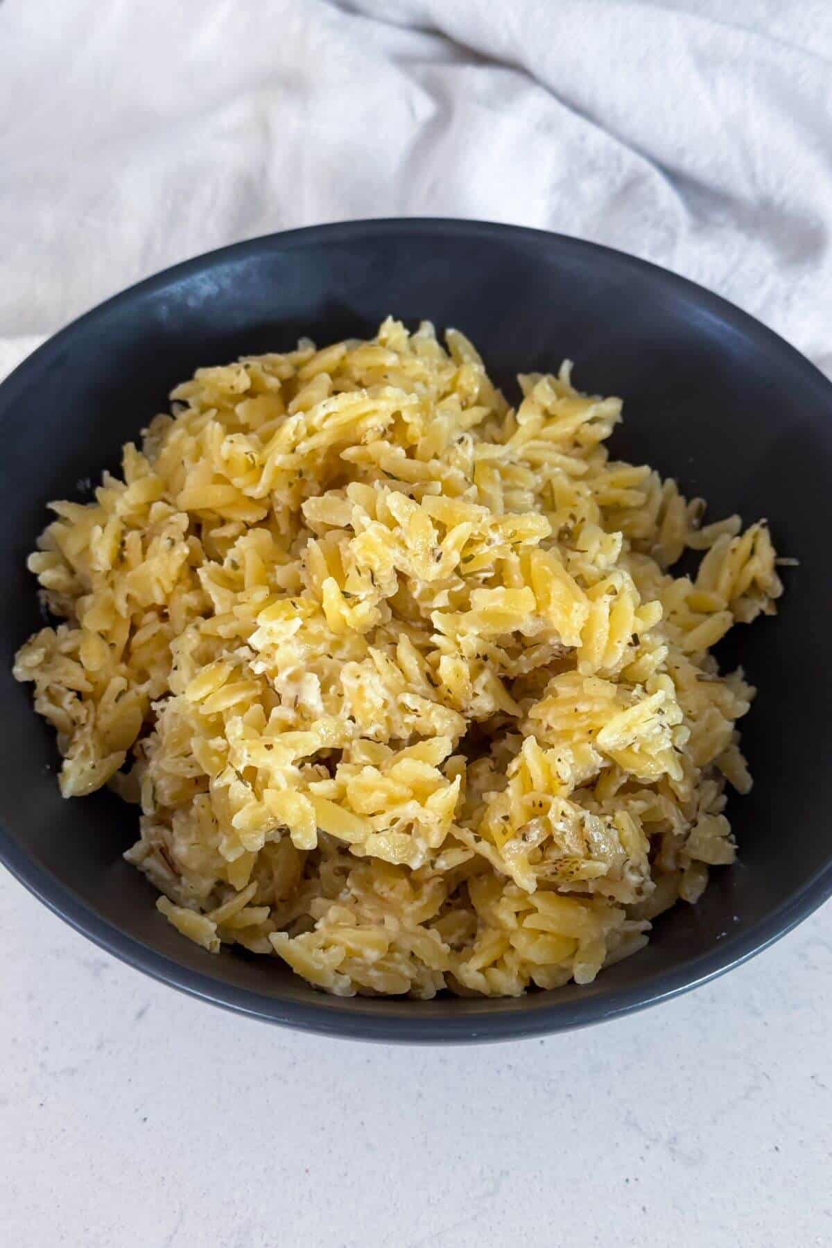 A black bowl filled with cooked orzo pasta mixed with herbs and grated cheese, placed on a white surface with a light fabric in the background.