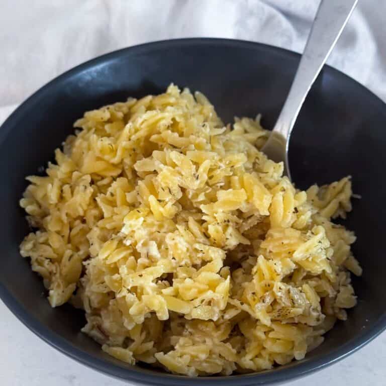 A black bowl filled with seasoned orzo pasta, with a fork resting inside, on a light surface.