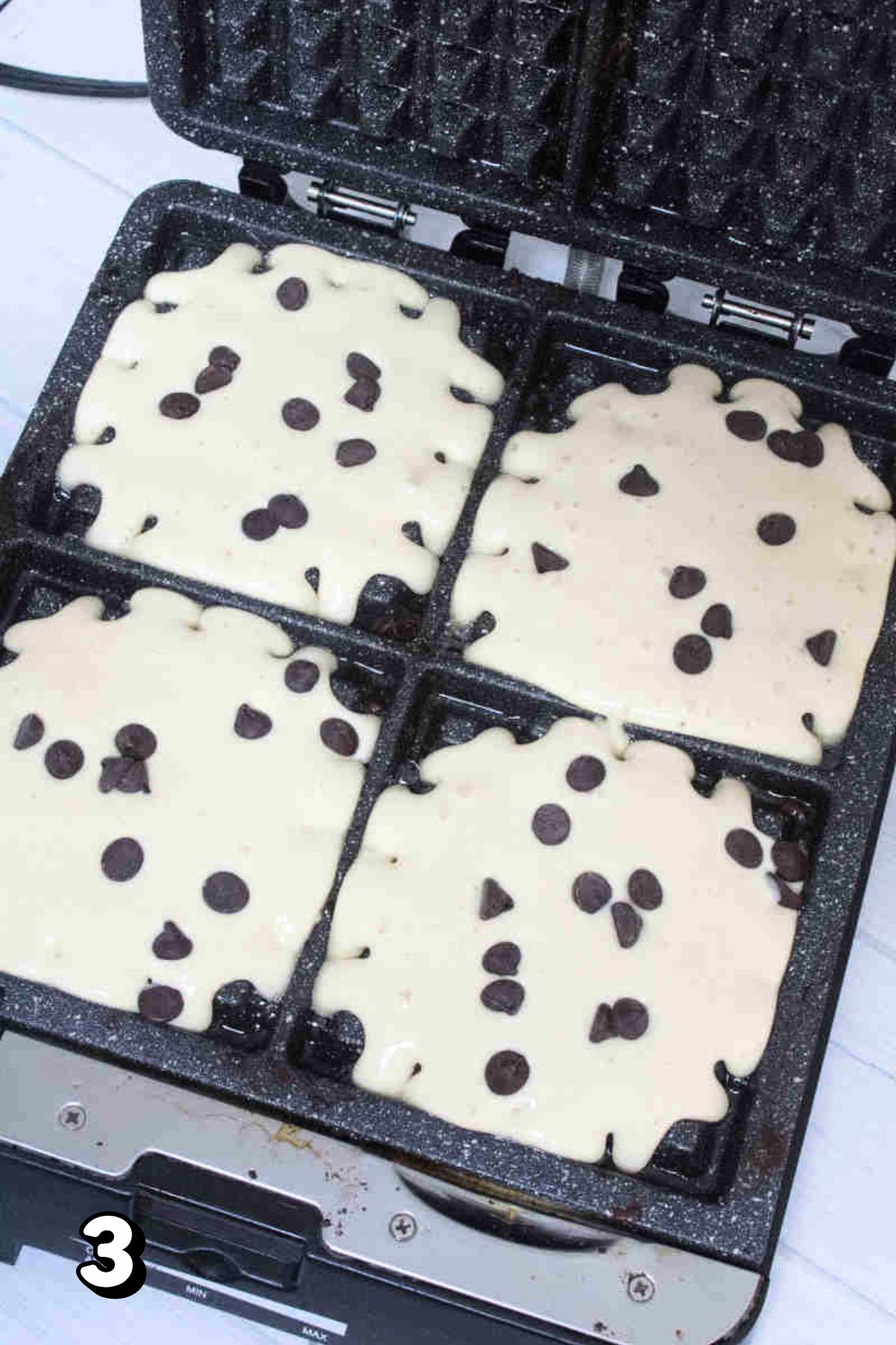 Raw waffle batter with chocolate chips in a waffle iron.