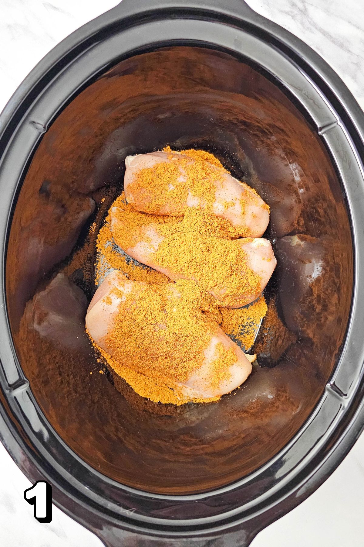 Raw chicken with taco seasoning on top in a black slow cooker.