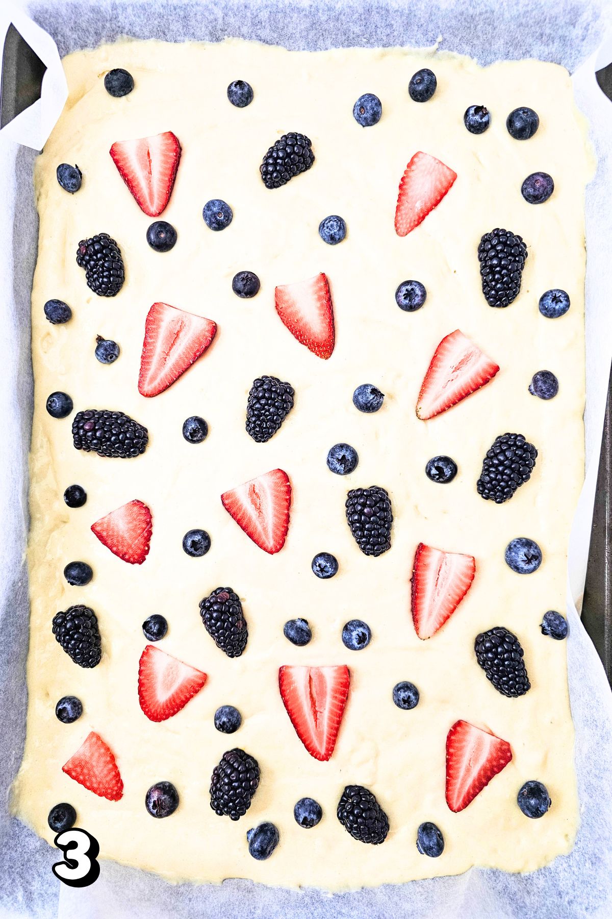 Raw sheet pan pancakes with berries in the batter on a lined baking tray.