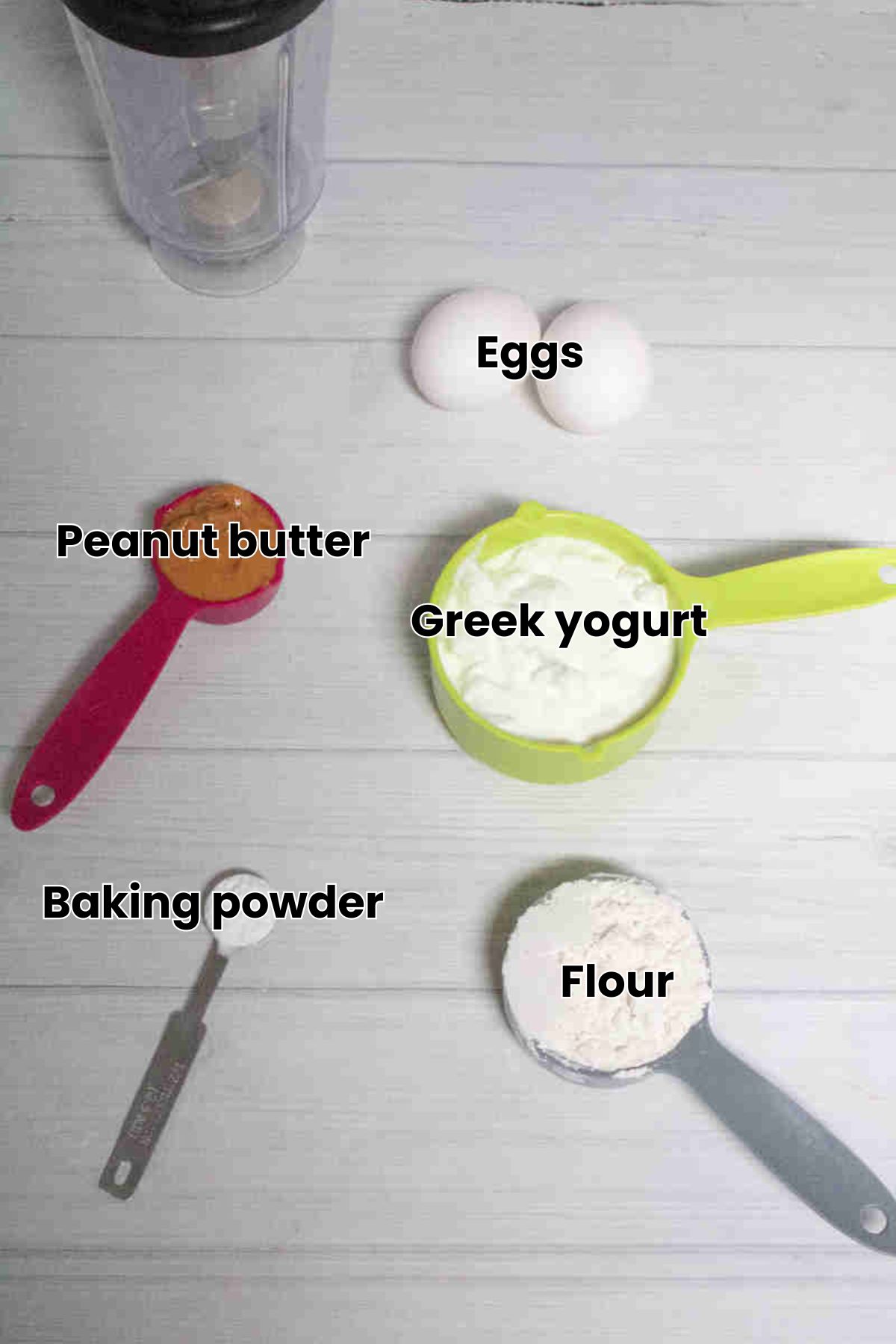 Greek yogurt, peanut butter, flour and more ingredients in measuring cups with text labels over the top./