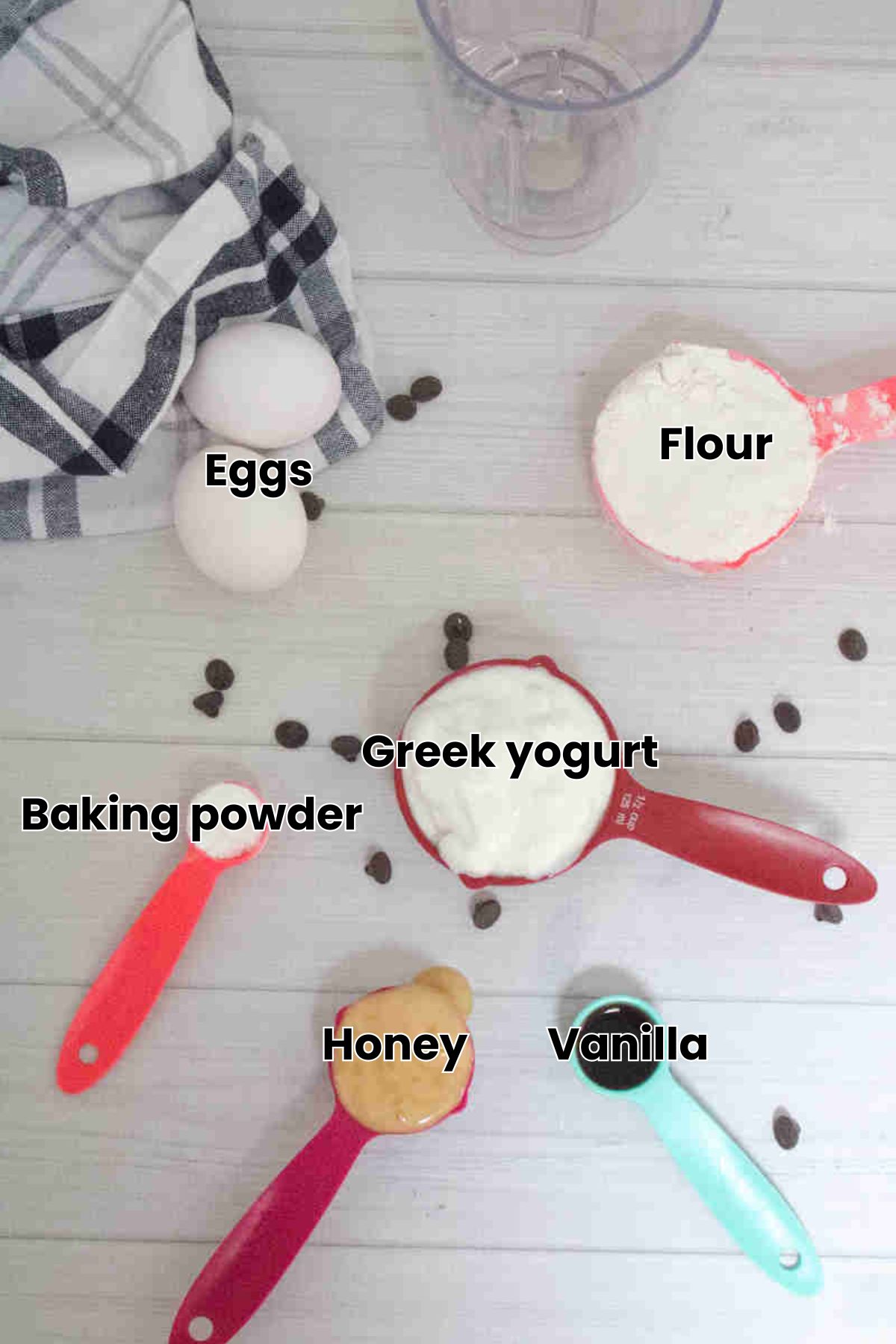 Multiple ingredients like yogurt, flours, eggs, and honey in measuring cups with labels over the top.