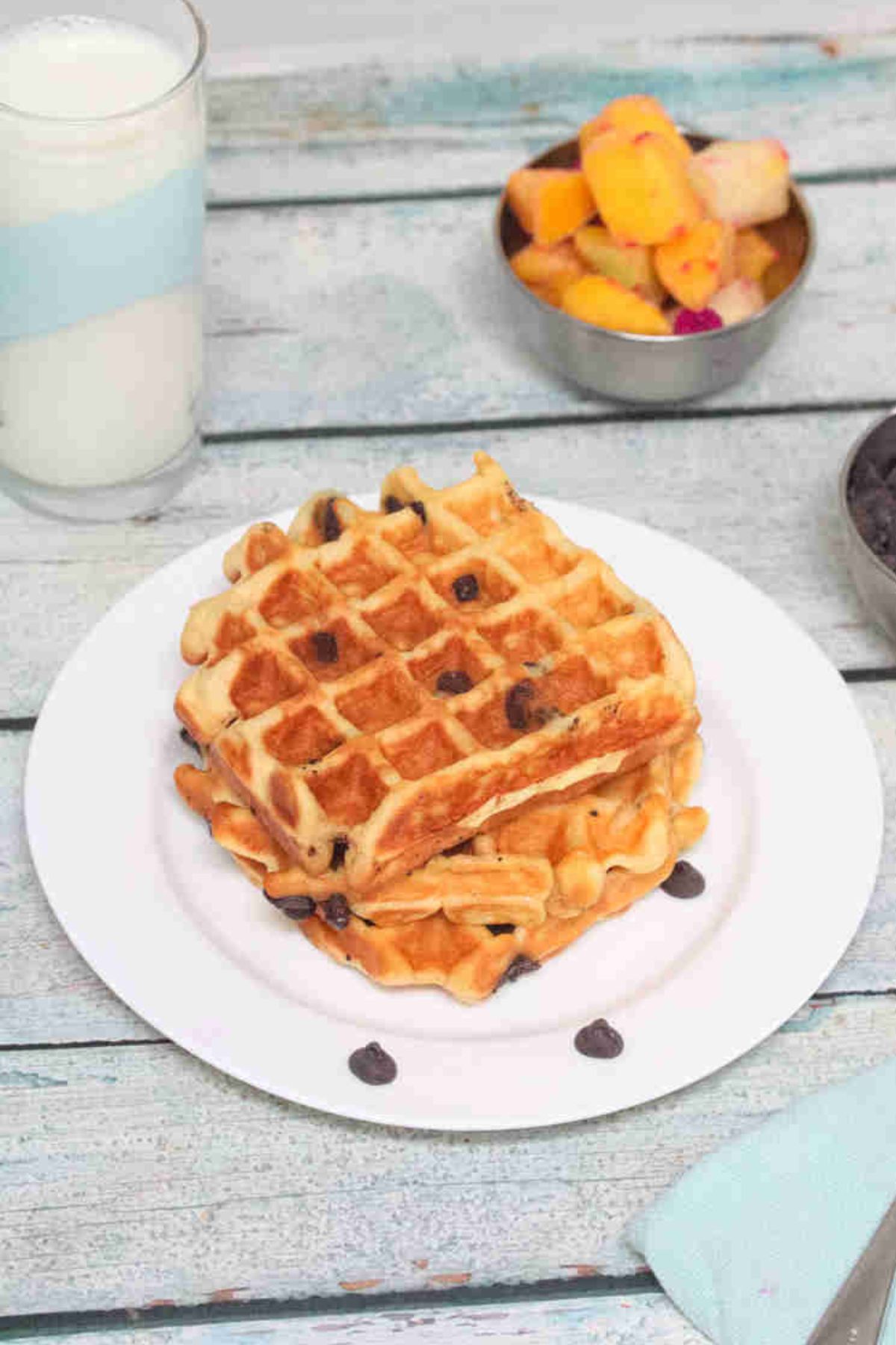 Three chocolate chip waffles stacked on a white plate with fruit behind them.