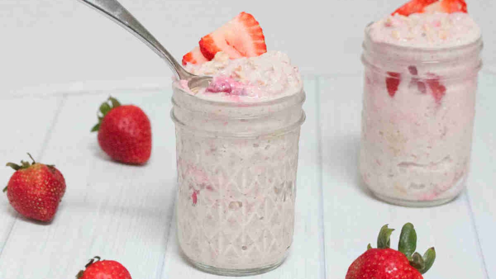 Two jars with blended yogurt overnight oats with strawberries and one jar has a spoon in it.