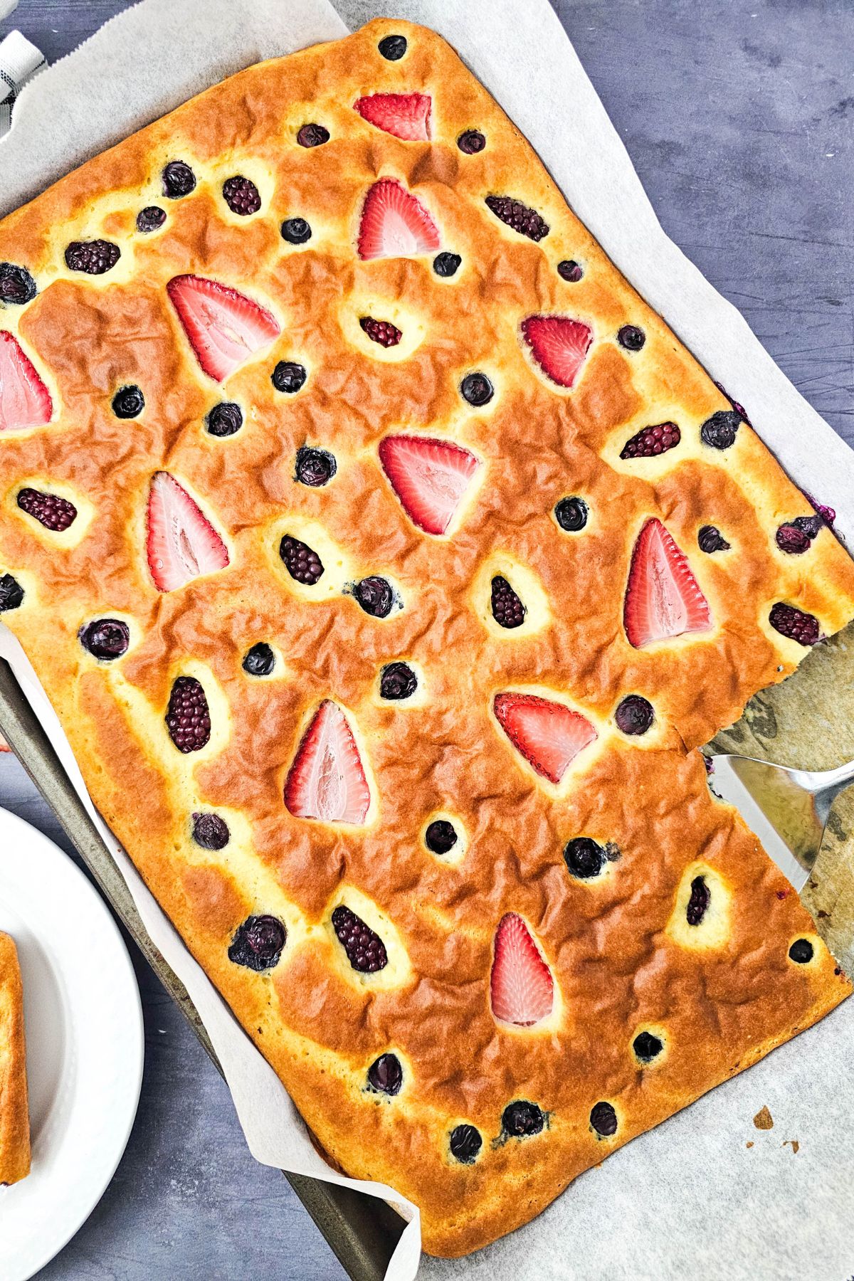 Sheet pan pancakes with berries on a baking sheet with one slice cut out.