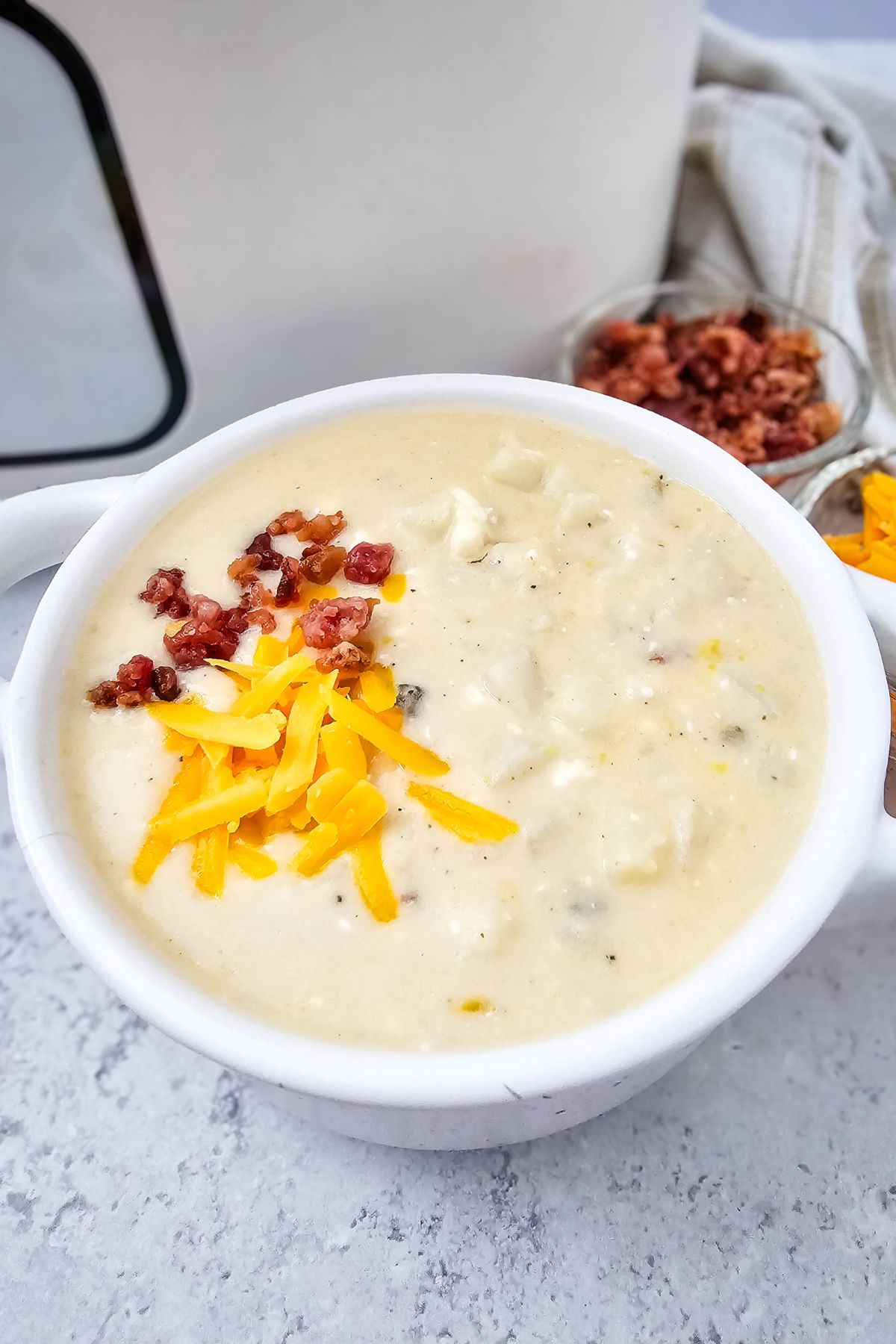 White bowl with loaded baked potato soup topped with cheese and bacon with a crockpot behind it.