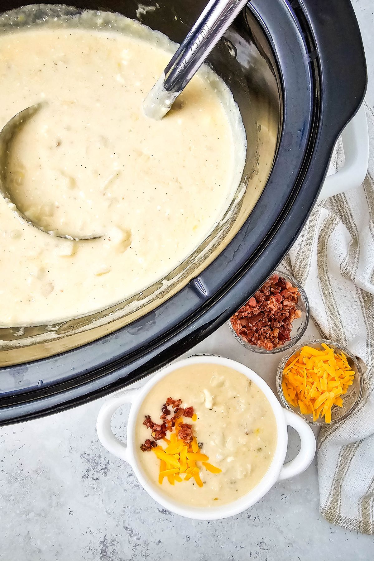 White bowl with loaded baked potato soup topped with cheese and bacon with a slow cooker behind it filled with soup.