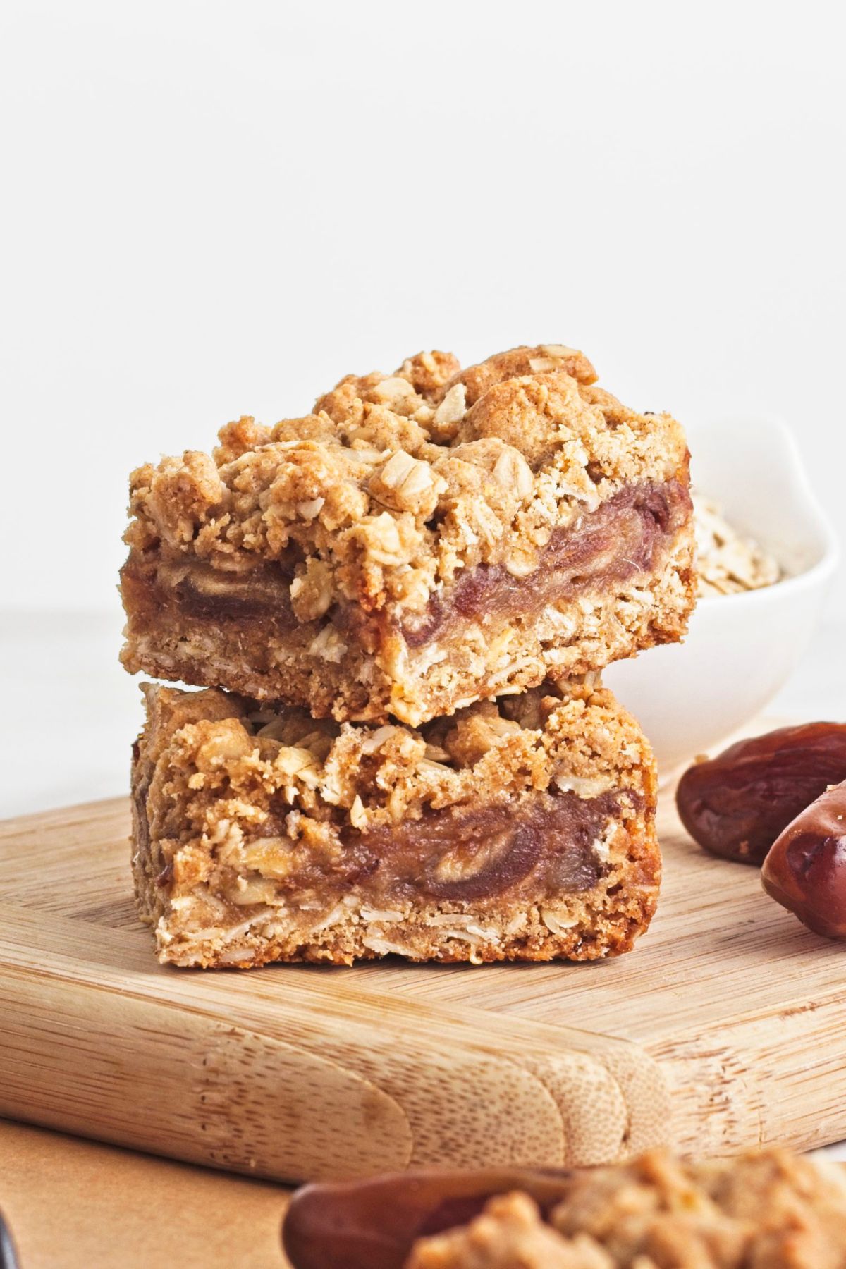 Two date and oat squares stacked on a cutting board with dates and more squares around them.