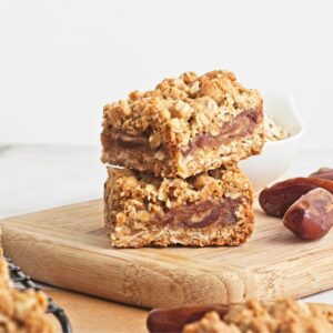 Two date and oat squares stacked on a cutting board with dates and more squares around them.