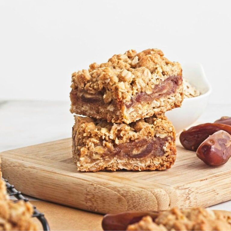 Two date and oat squares stacked on a cutting board with dates and more squares around them.