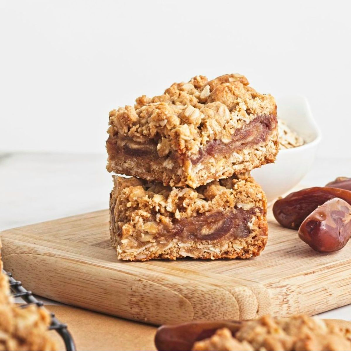Two date and oat squares stacked on a cutting board with dates and more squares around them.
