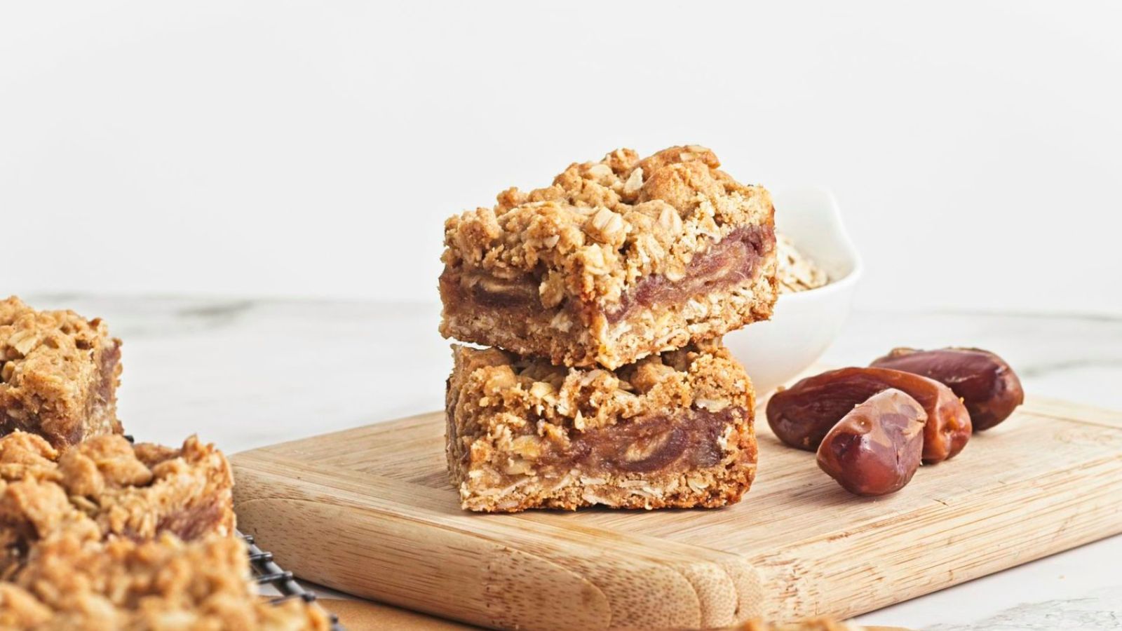 Two date and oat squares stacked on a cutting board with dates and more squares around them.