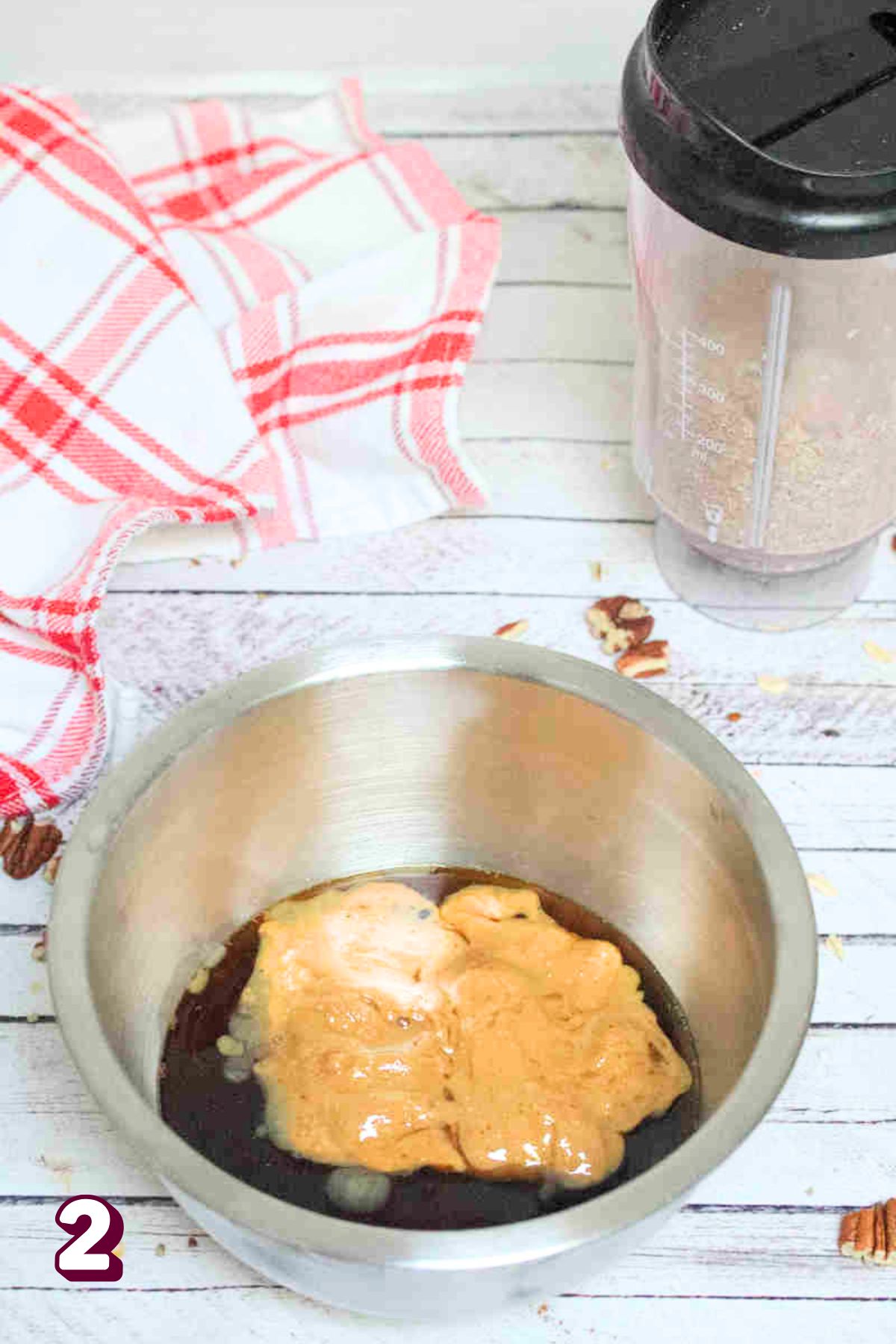 Steel bowl with almond butter, maple syrup, and vanilla in it with a mini blender behind it.