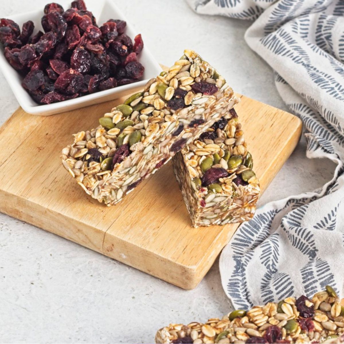 Two granola bars without peanut butter stacked on a wooden cutting board with dried cranberries behind it.