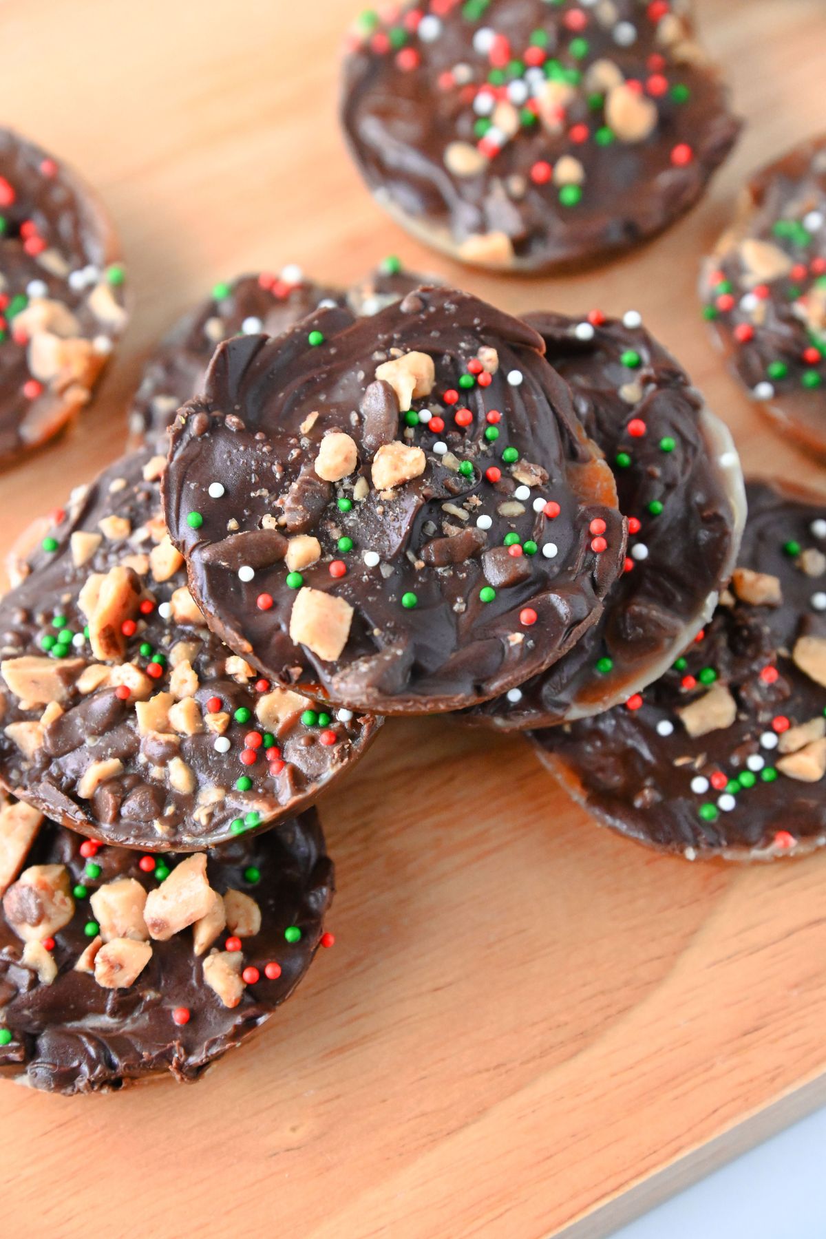 Multiple Christmas crack cookies topped with chocolate, toffee, and sprinkles, stacked on a wooden cutting board.